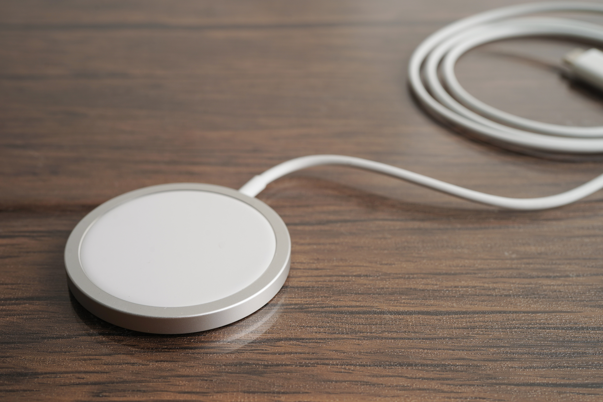 A sleek silver MagSafe charger is captured against a minimalist white background. The charger's circular design features a magnetic attachment point in the center, intended for effortless and secure charging of compatible Apple devices. The charger is connected to a white USB-C cable that trails off the frame, symbolizing its connection to a power source