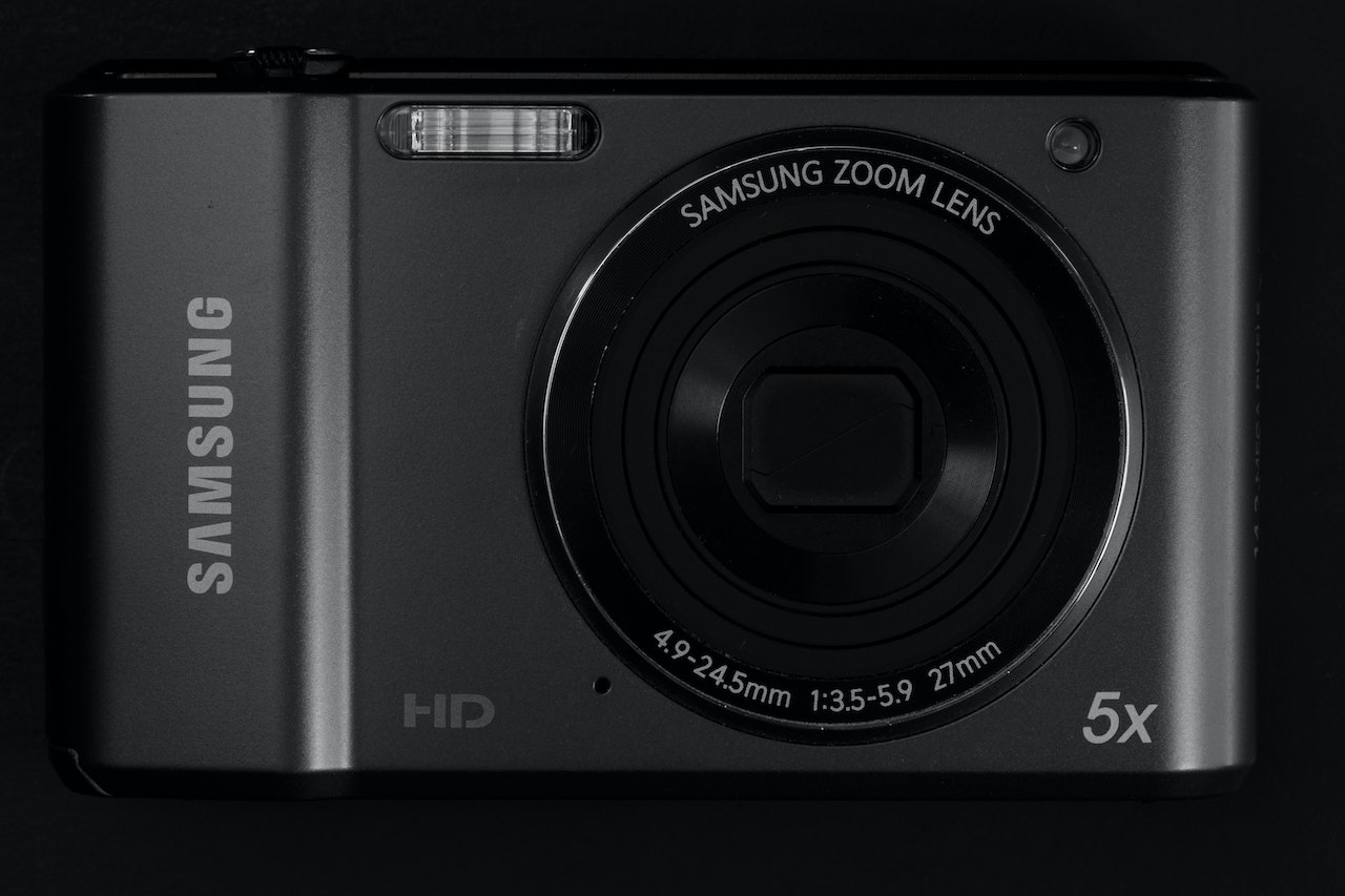 A sleek black Samsung camera, featuring a flip-up touchscreen display, is positioned on a white surface with its lens facing outward. The Samsung logo is clearly visible on the front of the camera, near the grip. The surrounding area shows photography accessories, including an SD card and lens cap, suggesting a professional or hobbyist setting