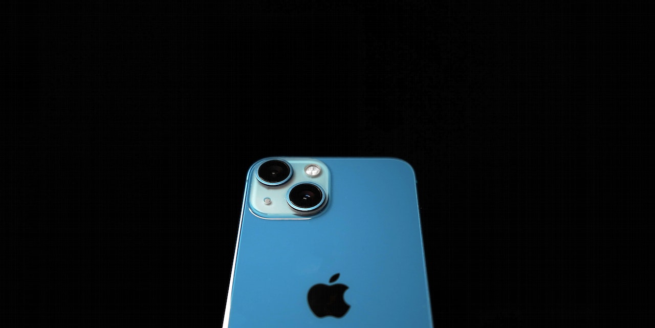 A sleek iPhone 13 showcased on a white table against a soft-focus background. The device is lying flat with the screen facing upward, displaying a vibrant wallpaper with a geometric design. The dual-camera system on the back is visible, demonstrating the phone's advanced photography capabilities. Ambient light casts a gentle glow on the phone, highlighting its premium glass finish and aluminum frame